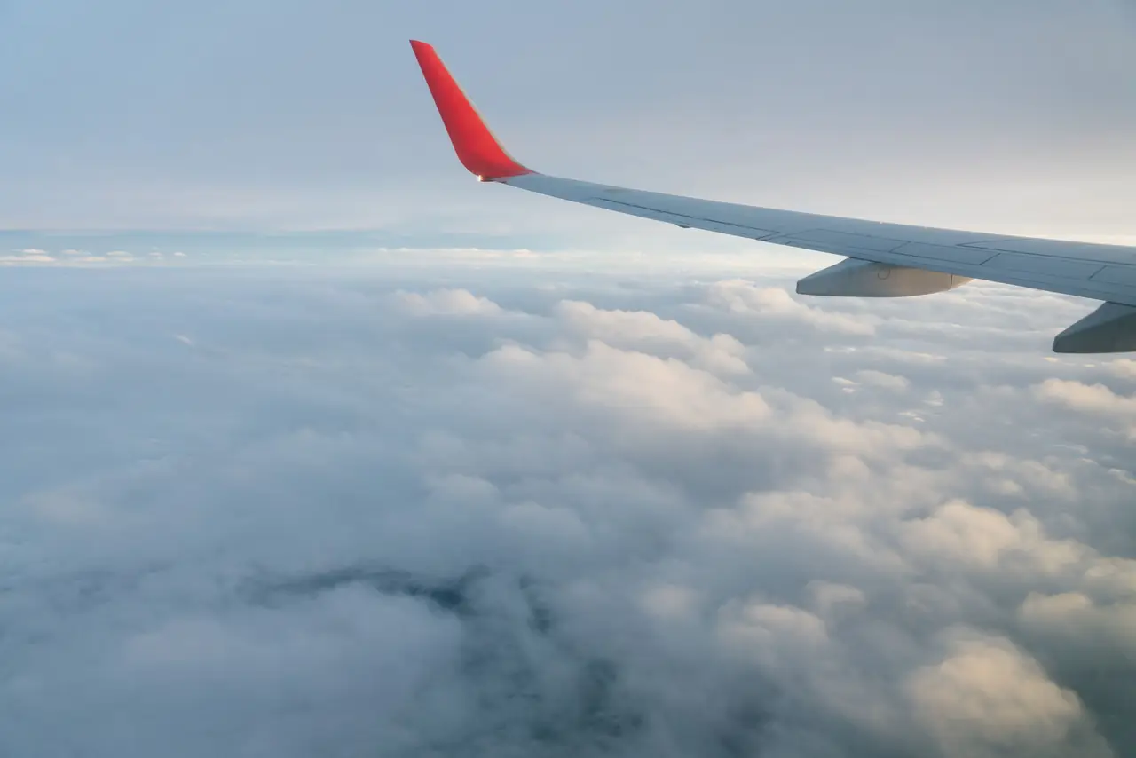 An airliner wing with a red winglet over a soft sea of clouds, seen from a window seat